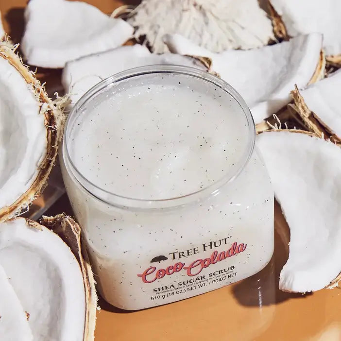 Jar of Tree Hut Coco Colada Shea Sugar Scrub surrounded by coconut pieces on a wooden surface.