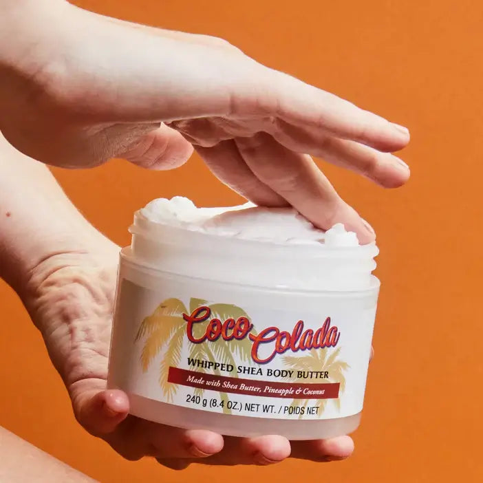 Tree Hut Coco Colada whipped shea body butter jar with a hand holding it against an orange background