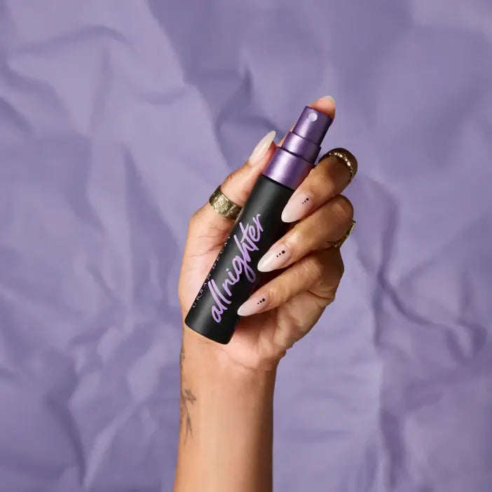 Hand holding a Urban Decay 'allnighter' spray bottle against a purple background
