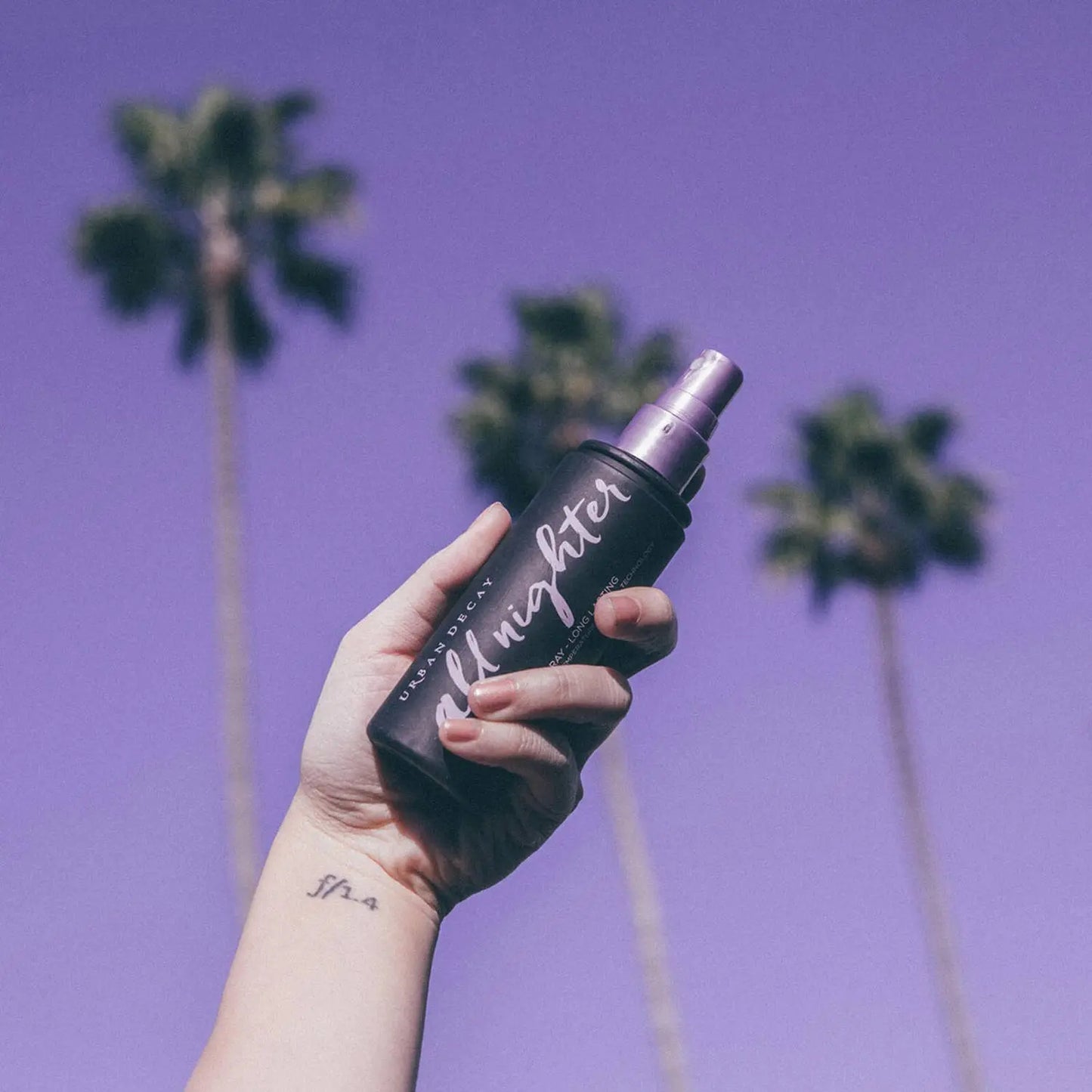 Urban Decay All Nighter Setting Spray held in hand against a purple sky with palm trees, perfect for long-lasting makeup.