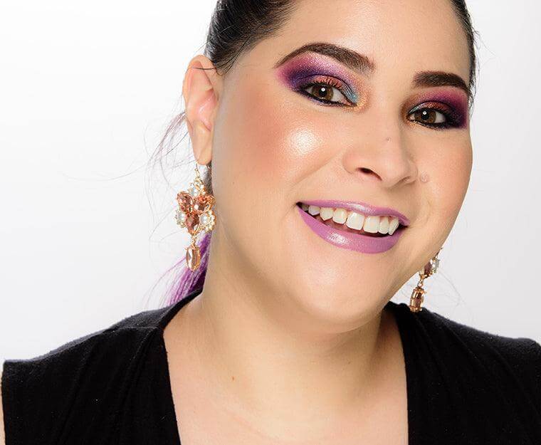 Woman with vibrant makeup featuring purple and blue eyeshadow, smiling brightly in elegant earrings.