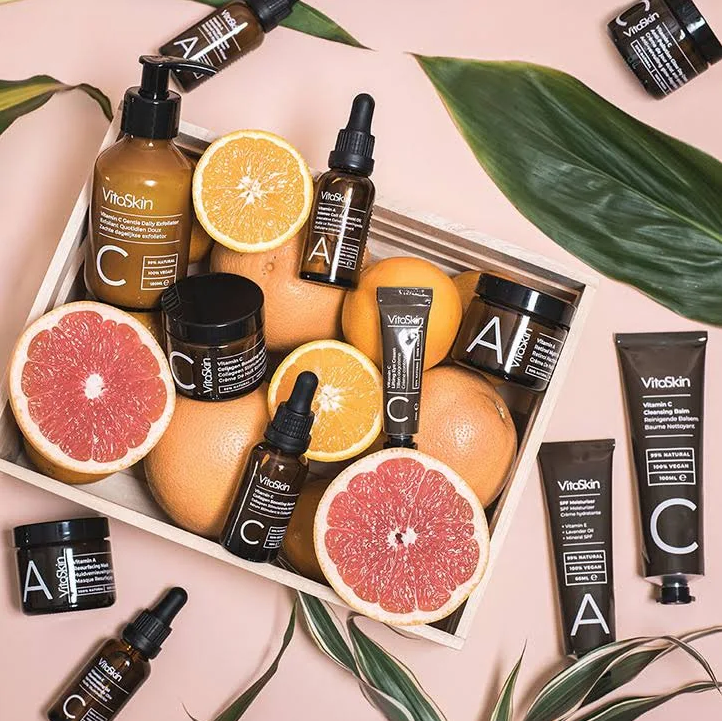 A variety of Vitaskin vitamin C skincare products surrounded by oranges and grapefruits in a wooden crate.