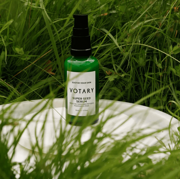 Votary Super Seed Serum bottle surrounded by lush green grass on a white plate.
