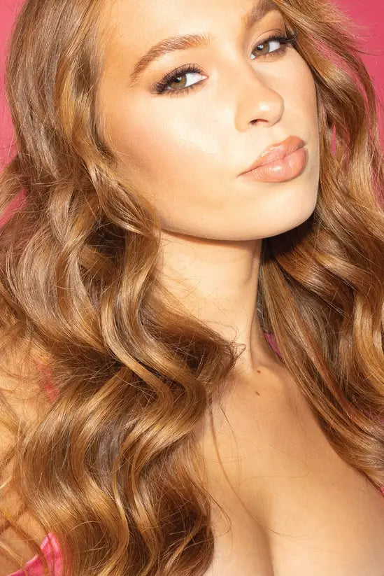 A close-up portrait of a woman with long, wavy light brown hair, wearing makeup and looking directly at the camera against a pink background.