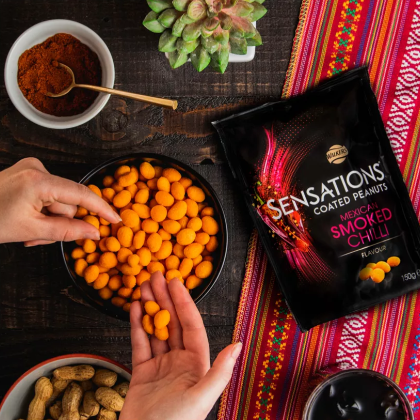Hands reaching for Sensations Mexican Smoked Chilli Coated Peanuts in a bowl on a wooden table.