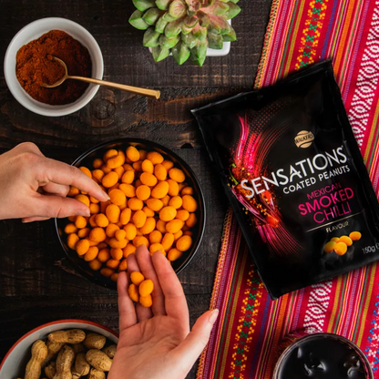 Hands reaching for Sensations Mexican Smoked Chilli Coated Peanuts in a bowl on a wooden table.