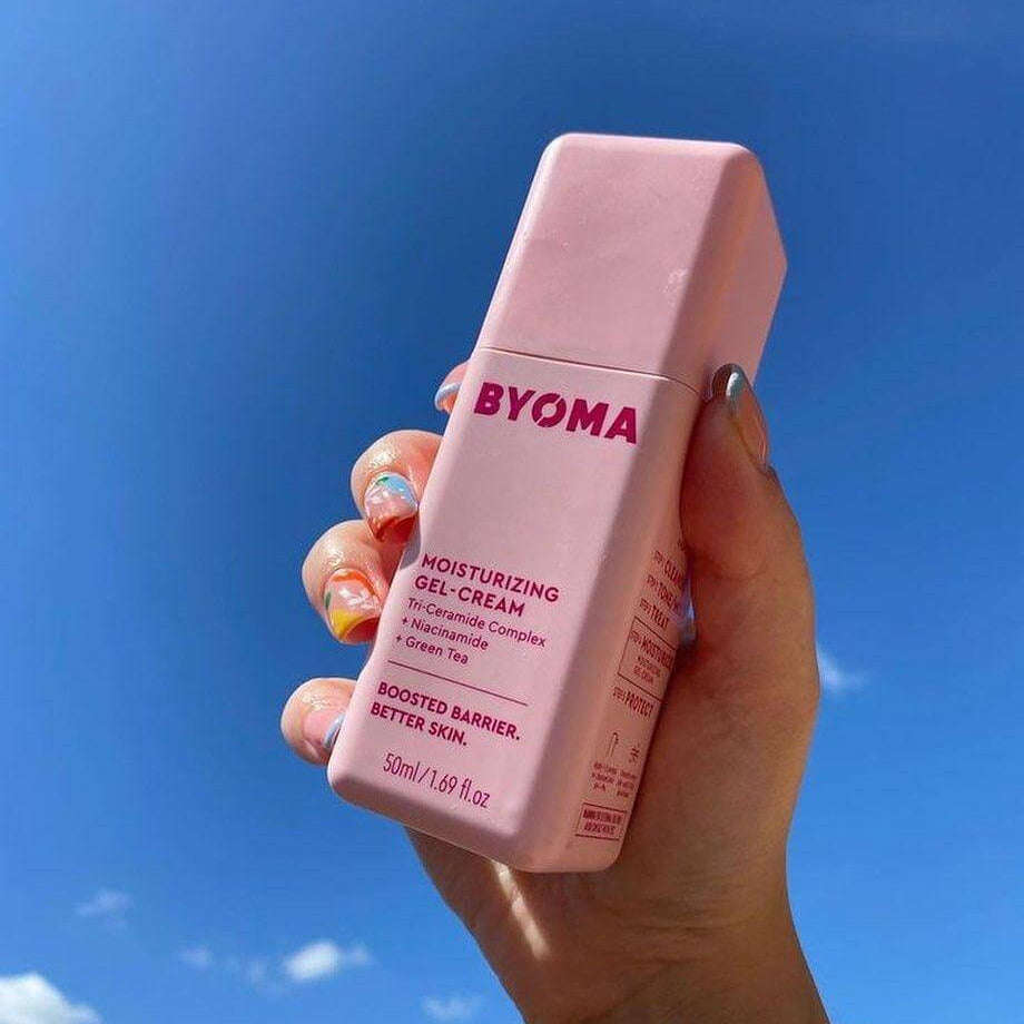Hand holding BYOMA Moisturising Gel Cream 50ml against a blue sky, showcasing its colorful packaging.