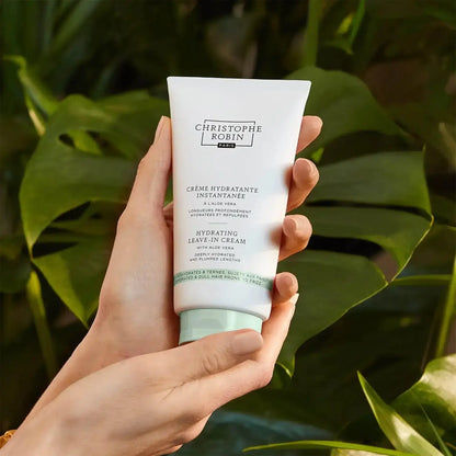 Hand holding CHRISTOPHE ROBIN Hydrating Leave-in Cream among green foliage, showcasing hydration for healthy hair.