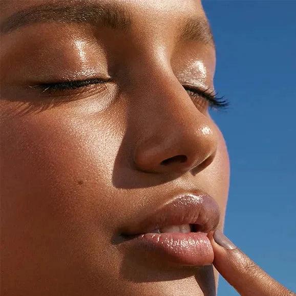 Close-up of a woman with Glossier glowing skin and soft lips, showcasing natural beauty with a blue sky background.