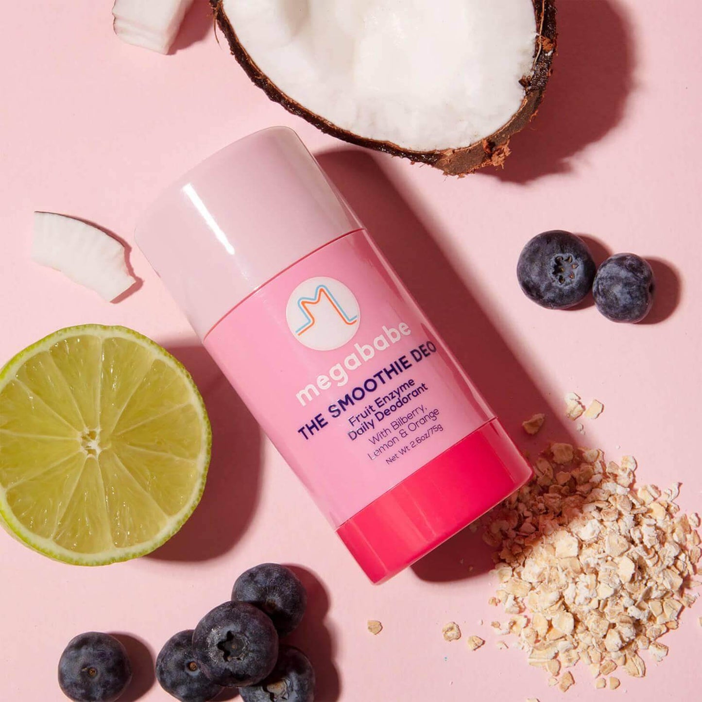 Megababe The Smoothie Deo fruit enzyme deodorant with lemon, coconut, and blueberries on a pink background.