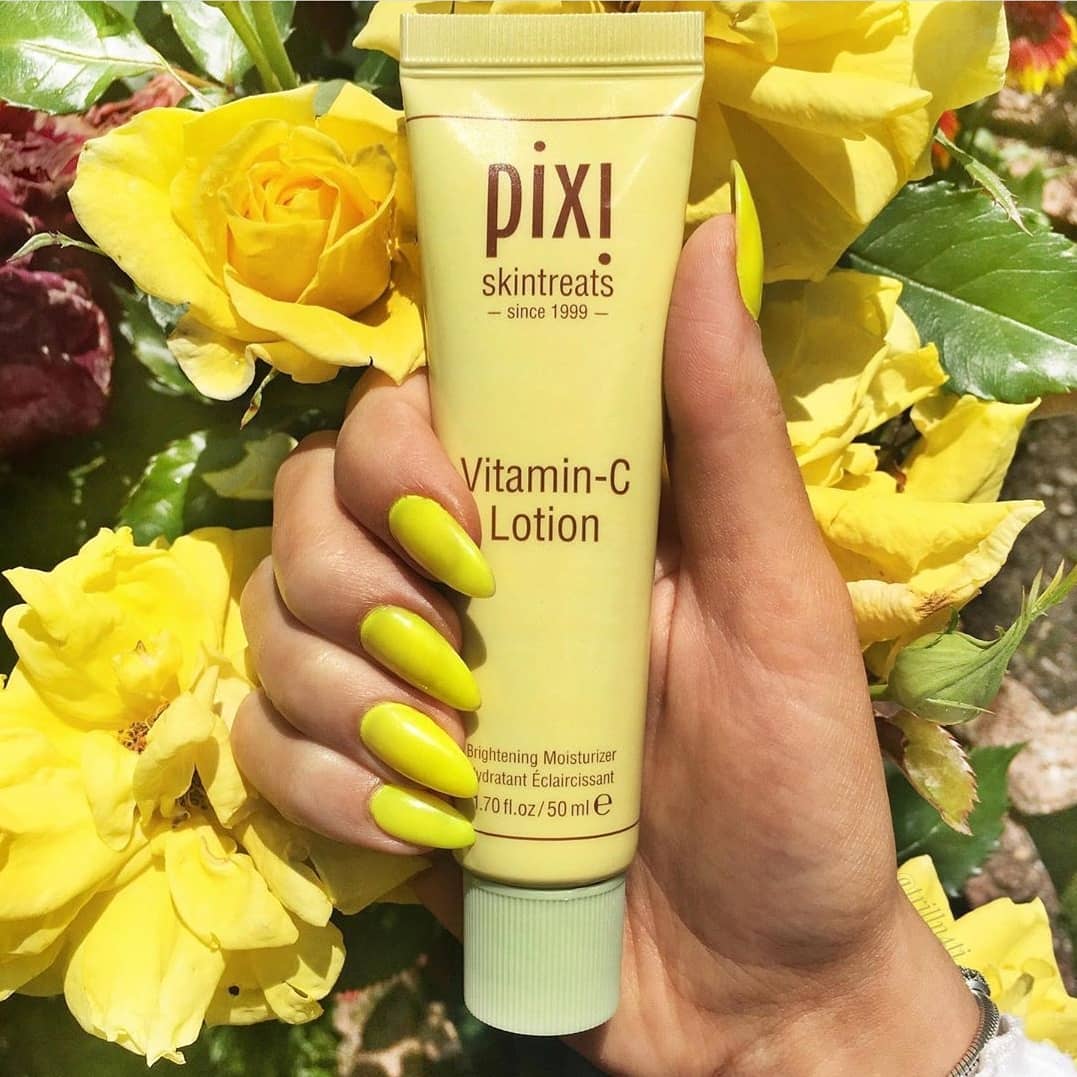 Pixi Vitamin-C Lotion 50ml held by hand with yellow flowers in the background, promoting skin brightness and radiance.