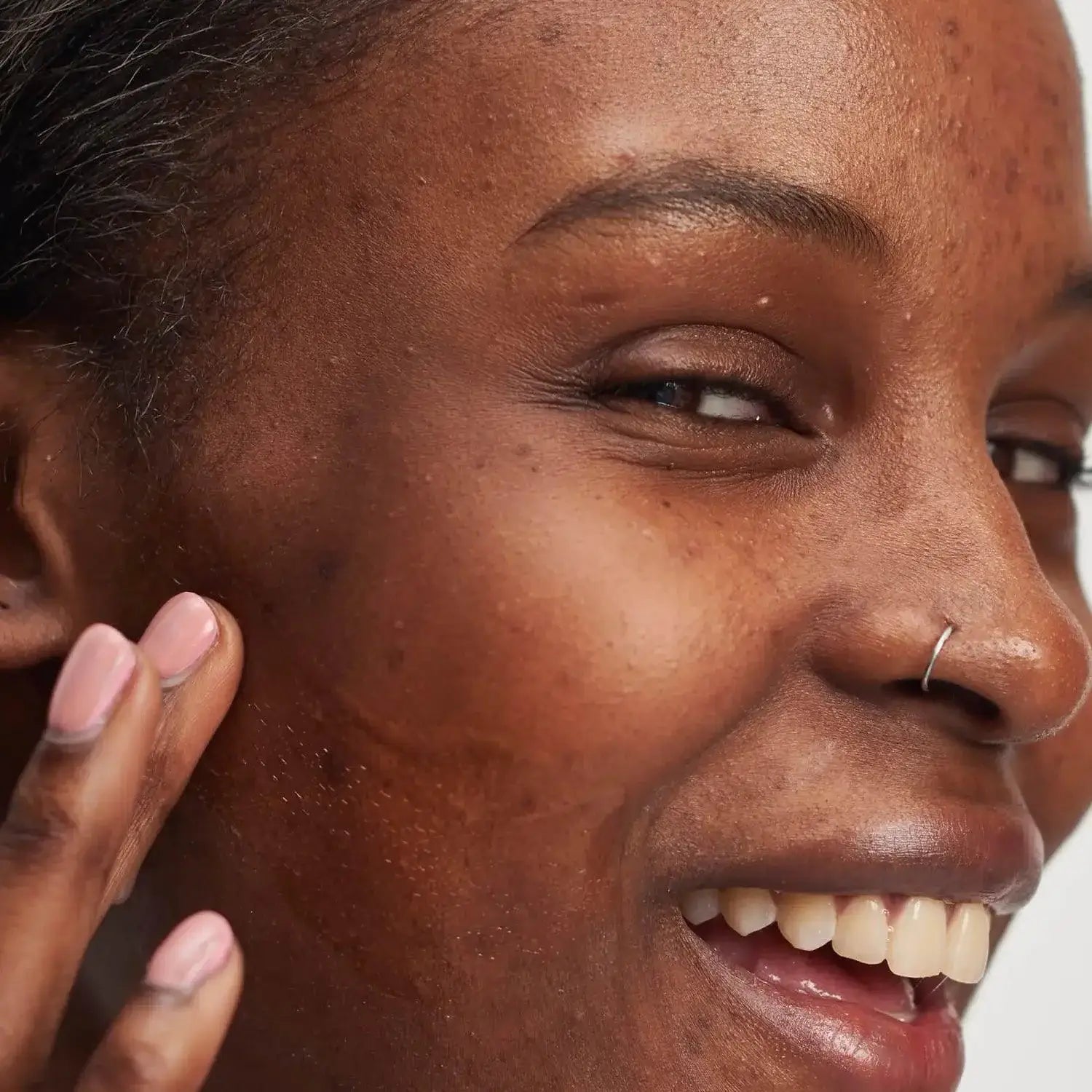Close-up of a woman smiling while applying skincare on her cheek, showcasing healthy glowing skin.